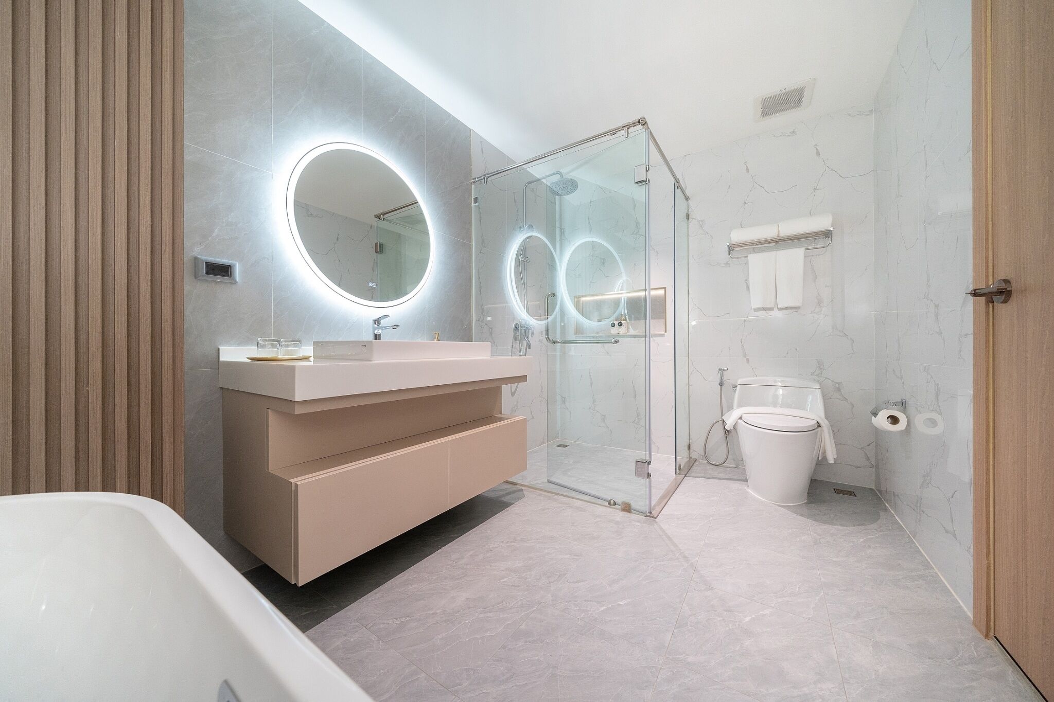A modern bathroom with a glass-enclosed shower, a round mirror with a light, a beige vanity, and a white toilet.