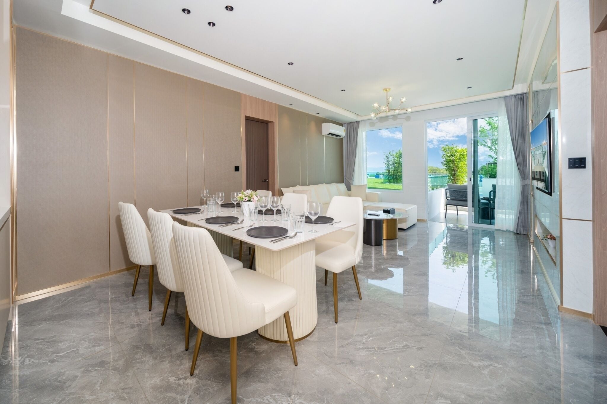 A modern dining area with a large table set for six, surrounded by white upholstered chairs with gold legs.