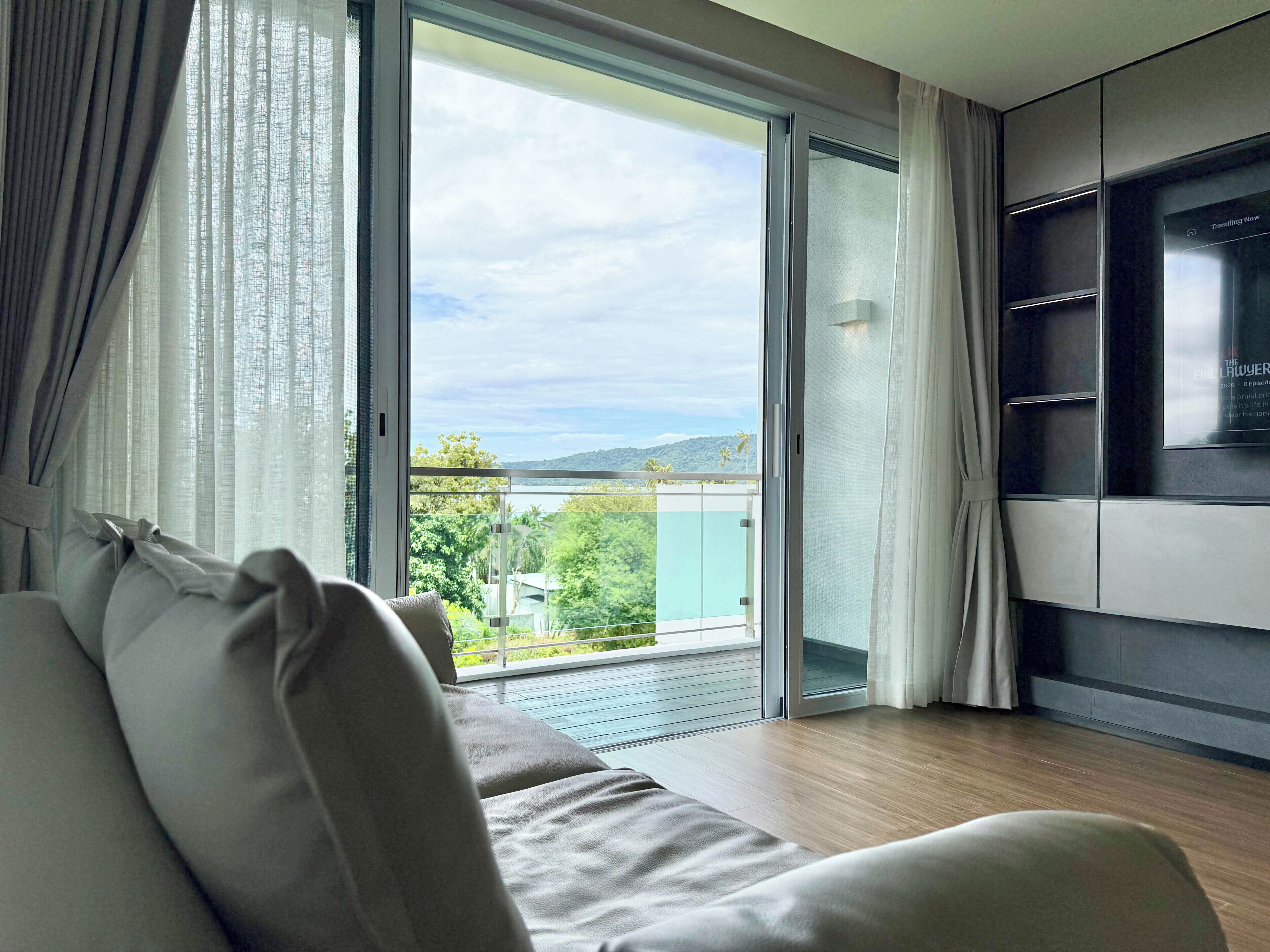 A modern living room with a sofa, a television, and a sliding glass door leading to a balcony with a view of greenery and hills.
