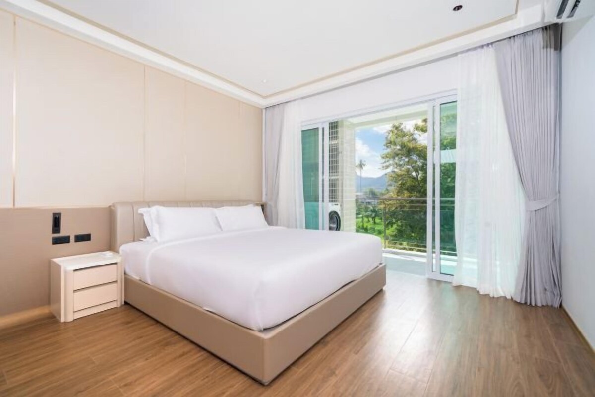 A modern hotel room with a large bed, a nightstand, and a view of a balcony with greenery.