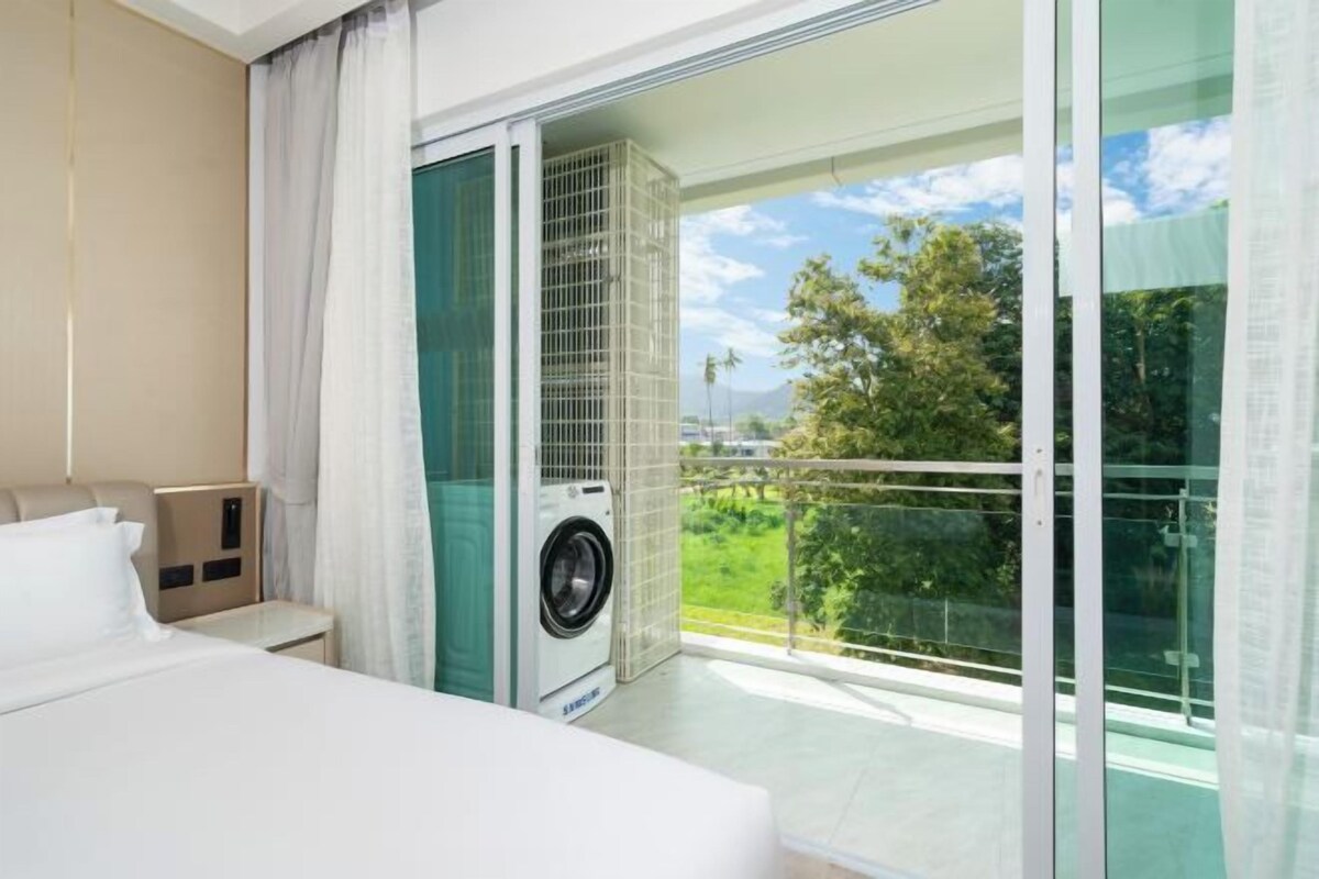 Habitación de hotel con cama, lavadora y balcón con vistas a zonas verdes y montañas.