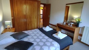 Un dormitorio con cama, dos almohadas, mesita de noche, cómoda, espejo y puerta que da a otra habitación.