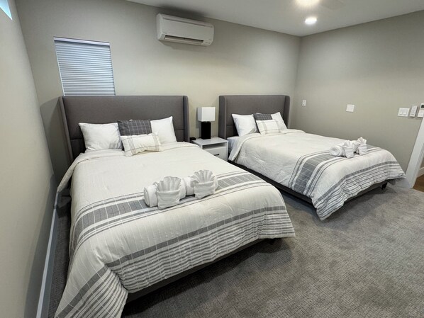 Habitación de hotel con dos camas, cada una con cabecero gris, ropa de cama blanca y una manta a rayas. Hay una mesita de noche con una lámpara, una ventana con persianas y un equipo de aire acondicionado en la pared.