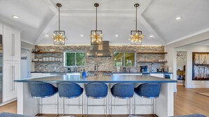 A modern kitchen with a central island, bar stools, pendant lighting, and a stone backsplash.