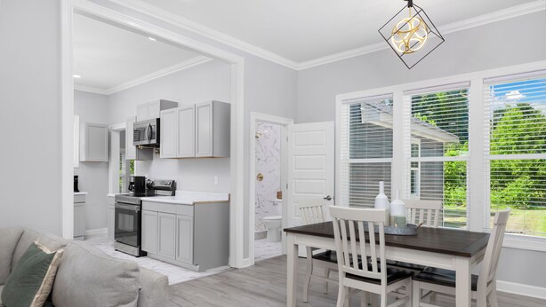 Une cuisine moderne avec des meubles blancs, un coin repas doté d’une table en bois foncé et de chaises blanches, et une grande fenêtre avec des stores.