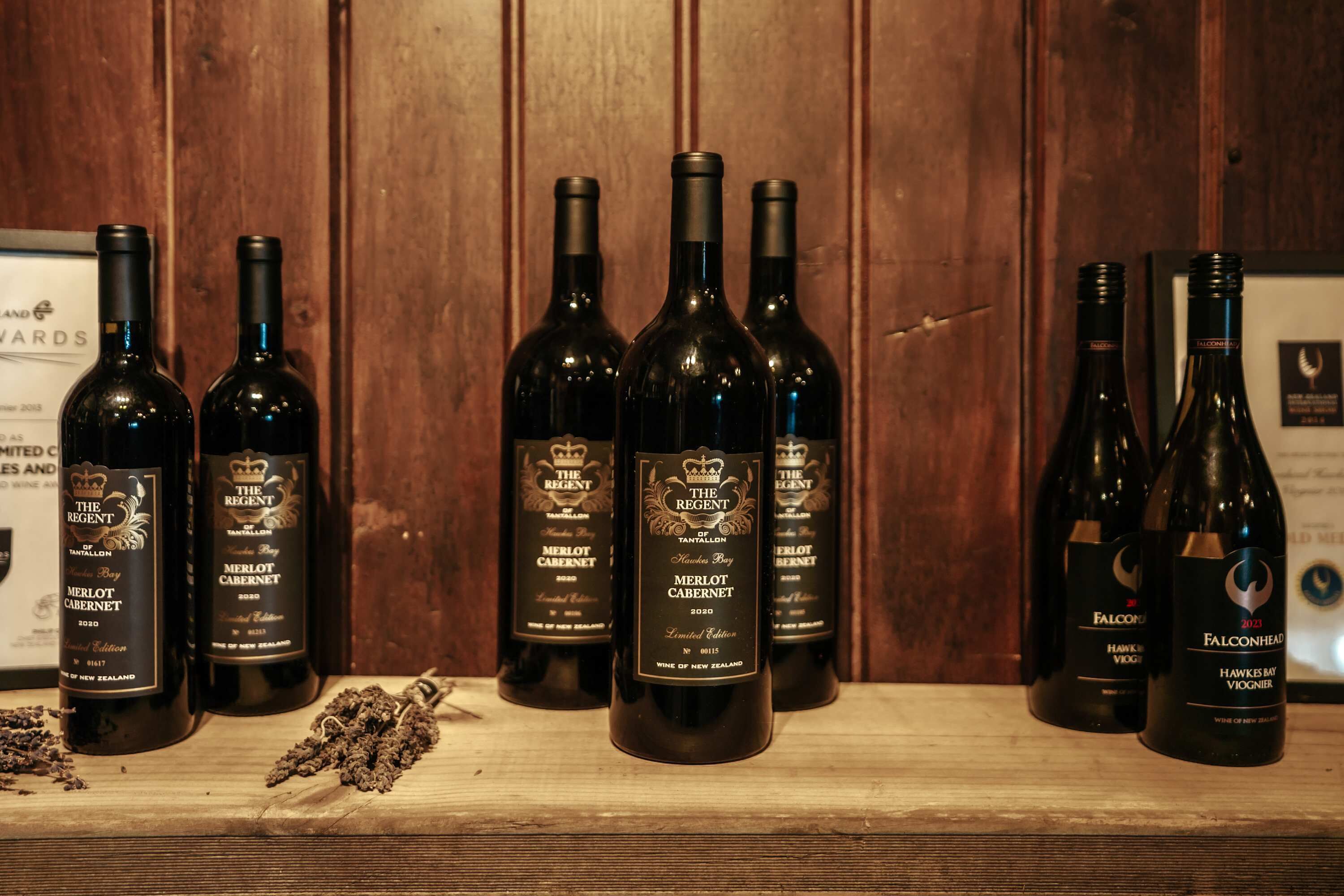 A wooden shelf with several bottles of wine, including 'The Regent Merlot Cabernet 2020' and 'Falconhead Hawke Bay Viognier 2020'.