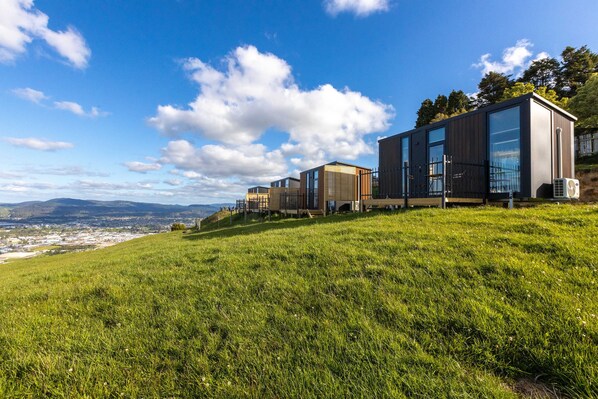 Un edificio moderno con grandes ventanales y techo plano, ubicado en una colina cubierta de hierba con vista a una ciudad distante.