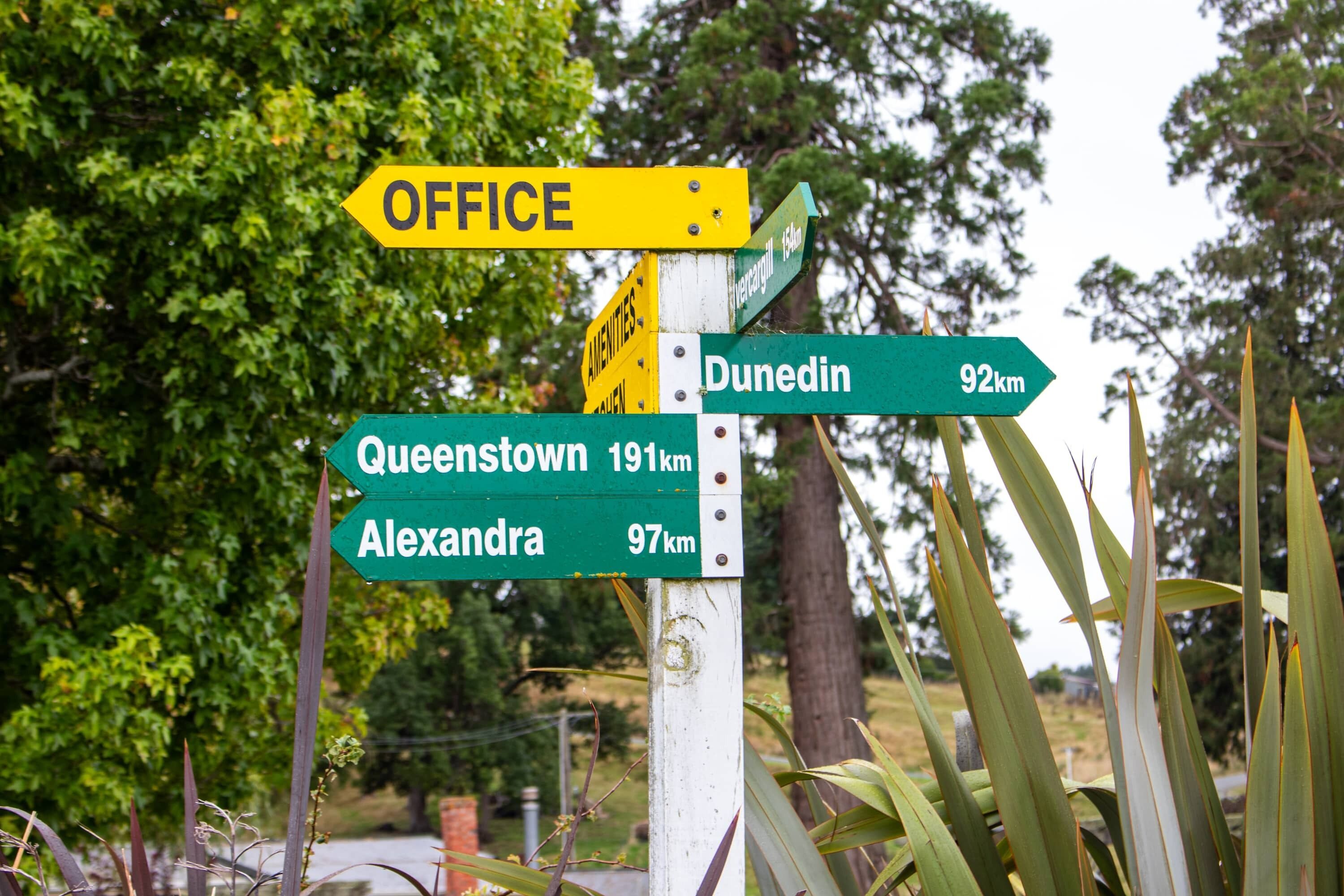 Un panneau de signalisation indiquant différentes directions, avec de nombreuses flèches pointant vers diverses destinations, notamment un bureau, Dunedin, Queenstown et Alexandra.