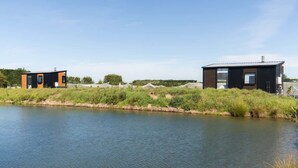 Two modern buildings by a calm water body, likely a canal, with a grassy embankment and trees in the background. - Vintners Retreat by Tiny Away (Burnham)
