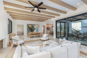 A modern living room with a ceiling fan, wooden beams, a dining area, and a view through large glass doors.