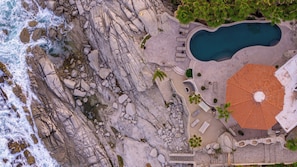 An aerial view of a coastal property with a swimming pool, outdoor seating, and a large circular patio.