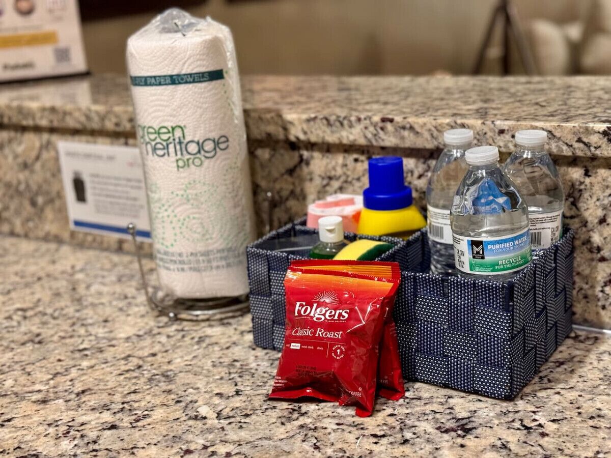 A countertop with a paper towel dispenser, a water bottle, a coffee packet, and a small basket containing various items.