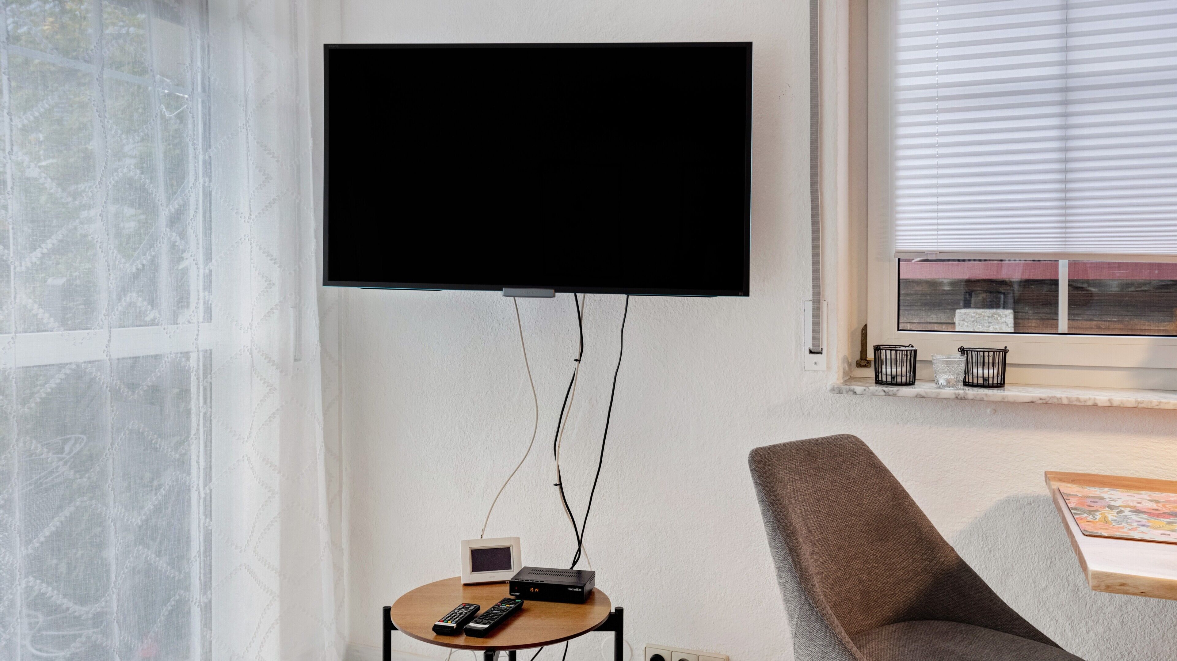 Ein Zimmer mit einem an der Wand befestigten Fernseher, einem kleinen Tisch mit Fernbedienung, einem Sessel und einem Fenster mit Jalousien.