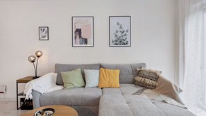 Een woonkamer met een grijze bank, een klein bijzettafeltje, een vloerlamp en drie ingelijste kunstwerken aan de muur.