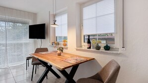 Spiseområde med et træbord, stole, et TV monteret på væggen og et vindue med persienner.
