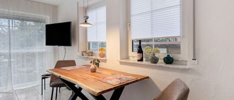 Un coin repas avec une table en bois, des chaises, un téléviseur fixé au mur et une fenêtre avec des stores.