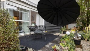 En terrasse med udendørs møbler, en stor parasol og en have med forskellige planter og pynteting.