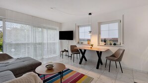 En moderne stue med sofa, sofabord, spisebord med stole, fjernsyn monteret på væggen og store vinduer med persienner.