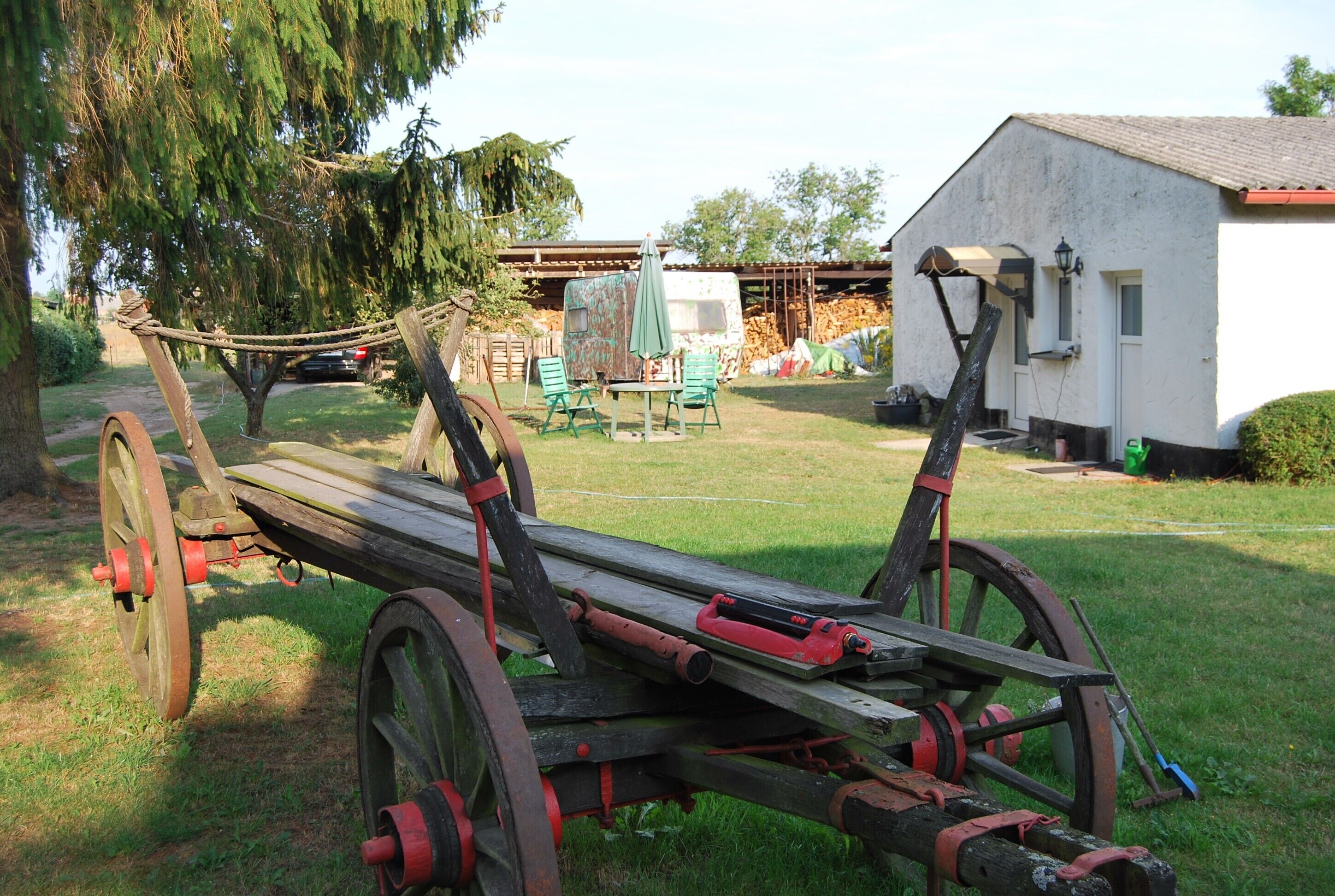Ein hölzerner Wagen mit Rädern und einer Holzbank in einer grünen Umgebung, im Hintergrund ein weißes Gebäude.