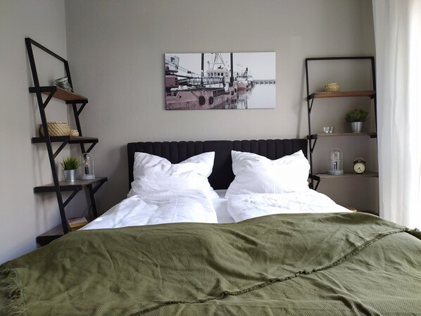 A neatly made bed with white linens, a green blanket, and a cityscape painting above it.