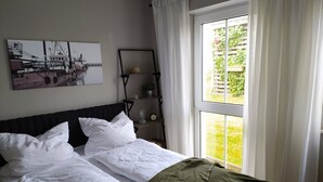 Un dormitorio con una cama, una ventana con cortinas, un estante en la pared con una planta y un cuadro.