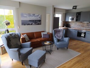 A modern living room with a sofa, two armchairs, a coffee table, and a kitchen area with appliances.