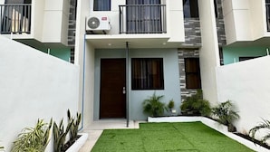 A modern two-story residential building with a flat roof, a central balcony, and a small front yard with artificial grass and potted plants.