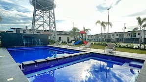 A swimming pool with a water tower labeled "MINA COAST" in the background, surrounded by lounge chairs and a small playground.