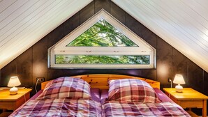 A bedroom with a sloped ceiling, a large window with a view of trees, a bed with plaid bedding, and two bedside lamps. - Apartment 'Ferienwohnung Klee 2' with Wi-Fi (WHV)