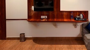 A room with a wooden TV stand, a mounted TV, a framed picture of roses, and a small shelf with a kettle and other items. - Plaza Hotel (Pithoragarh)