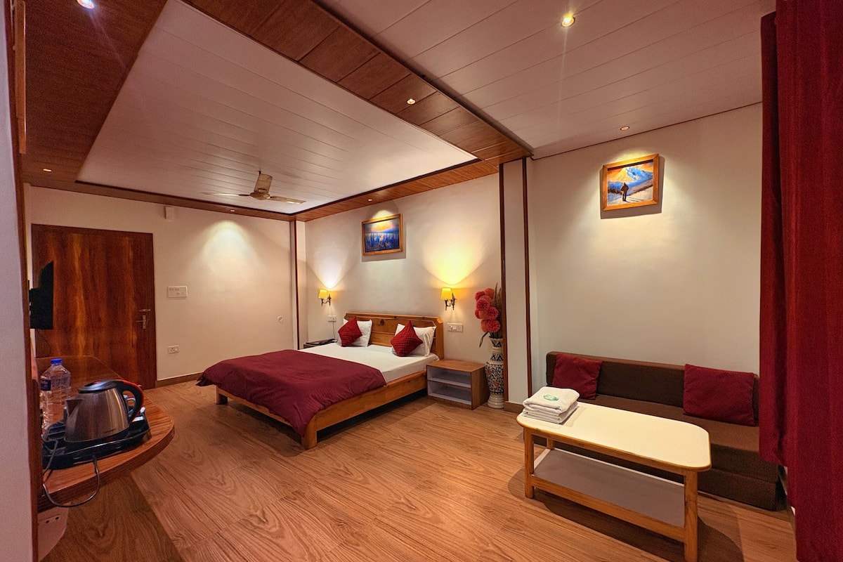 A hotel room with a wooden ceiling, a bed with red bedding, a bedside table, a sofa, and a painting on the wall.