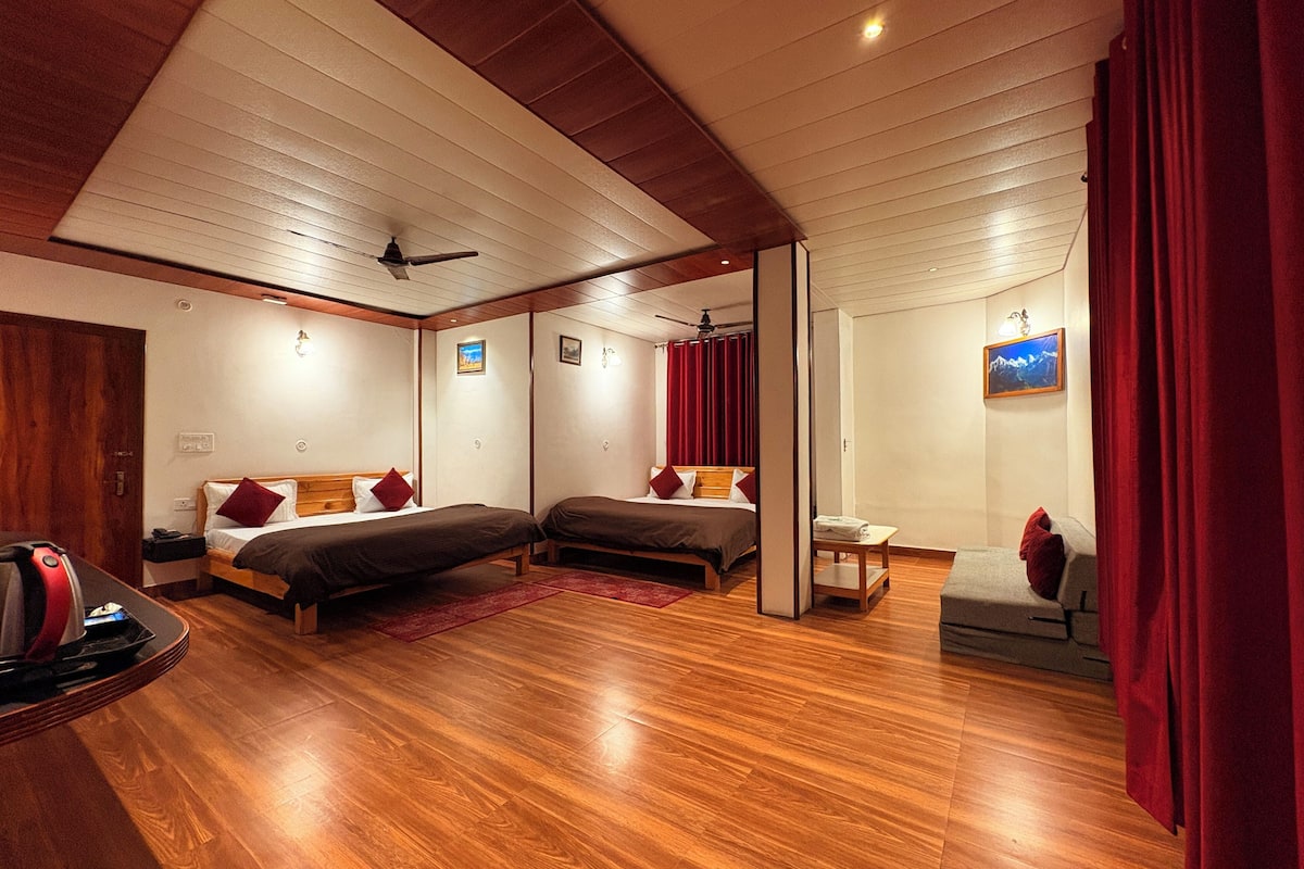 A hotel room with two beds, wooden flooring, ceiling fans, and a small table with a chair.