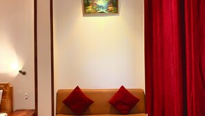 A room with a wooden ceiling, a beige sofa with red cushions, a small wooden table with a folded white towel, and a painting of a landscape on the wall. - Plaza Hotel (Pithoragarh)