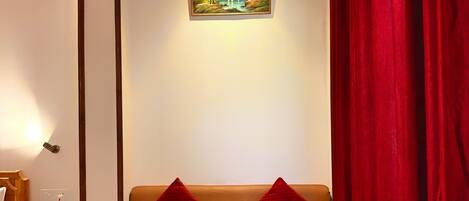 A room with a wooden ceiling, a beige sofa with red cushions, a small wooden table with a folded white towel, and a painting of a landscape on the wall.