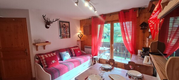 A cozy room with a wooden sofa, a dining table set for two, and a window with red curtains.