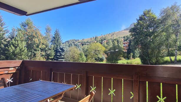 Una terraza de madera con una mesa y sillas, con vistas a un paisaje montañoso con árboles y edificios.