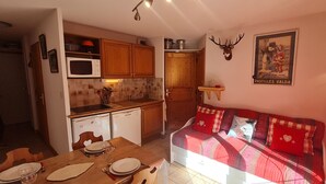 Cocina compacta con armarios de madera, microondas y lavavajillas. Hay una zona de comedor con una mesa preparada para dos personas y un sofá rojo con cojines estampados.