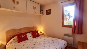 Un dormitorio con cabecera de madera, cama con ropa de cama floral, ventana con cortinas, radiador y estante con libros y un cuadro.