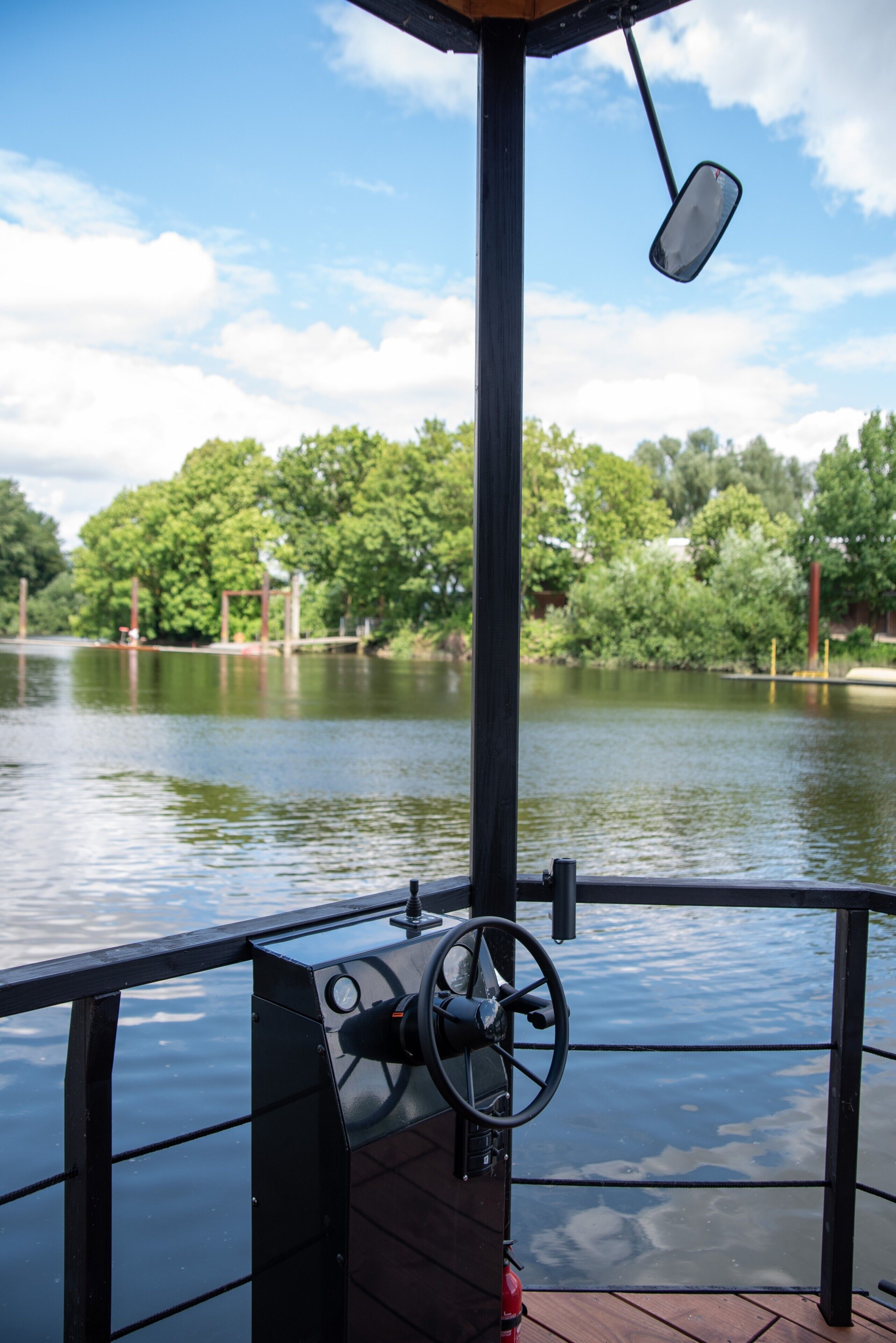 Ein Bootsdeck mit Steuer, Rückspiegel und Blick auf einen stillen See mit Bäumen im Hintergrund.