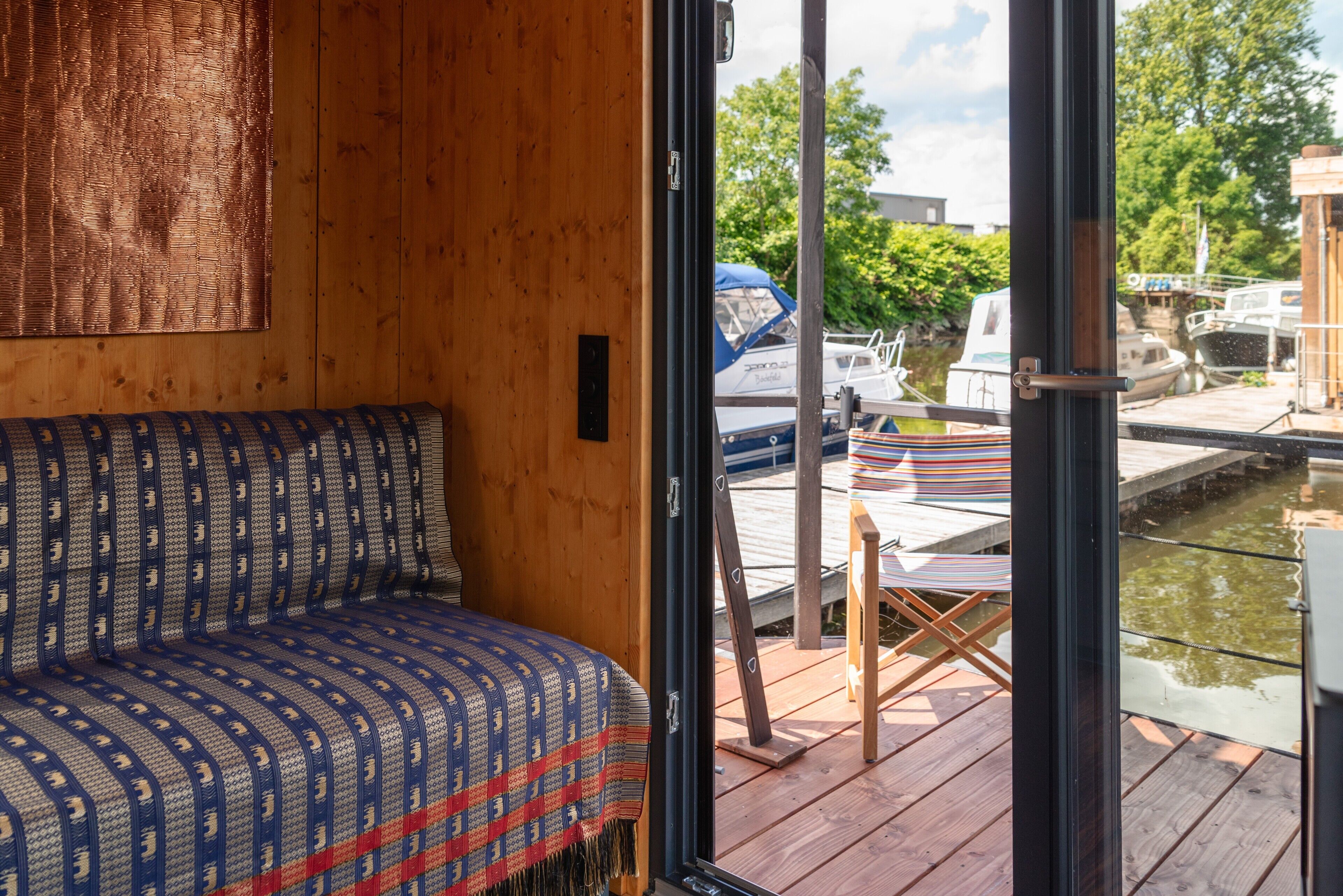 A wooden interior with a patterned bench, a glass door leading to an outdoor deck with a table and chairs, and a view of boats on a waterway.