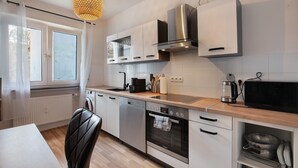 Eine moderne Küche mit Edelstahlgeräten, einem Geschirrspüler, einem Backofen und einer Mikrowelle. Ein Fenster mit durchsichtigen Vorhängen, eine Pendelleuchte und eine Holz Arbeitsplatte sind ebenfalls vorhanden.