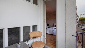 Ein Balkon mit Stuhl und Tisch, ein Schlafzimmer mit Bett und Nachttisch sowie Blick auf die Straße mit parkenden Autos.