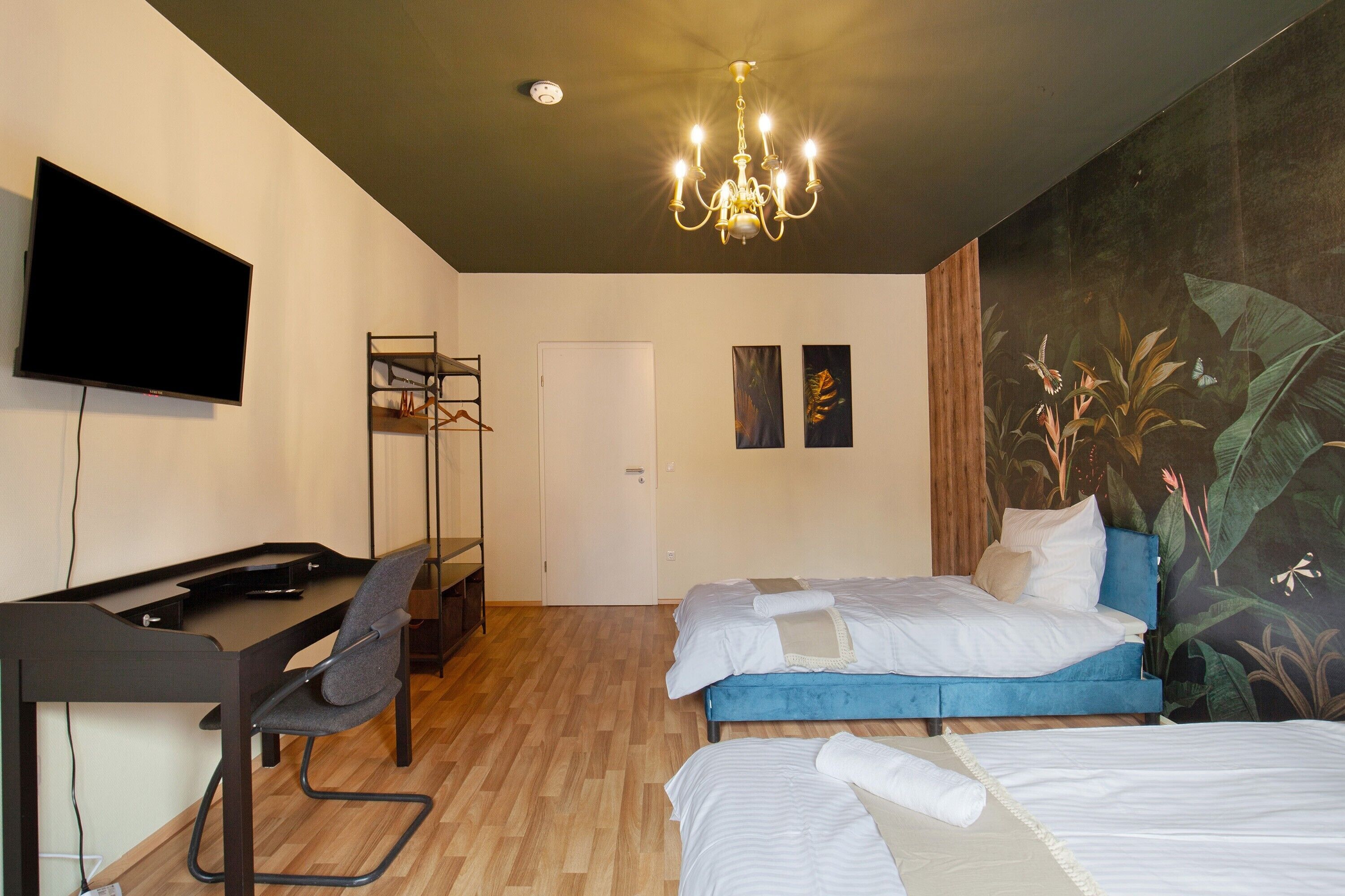 Une chambre d’hôtel équipée d’un téléviseur à écran plat, d’un bureau avec une chaise, de deux lits, d’un lustre et d’une fresque murale.