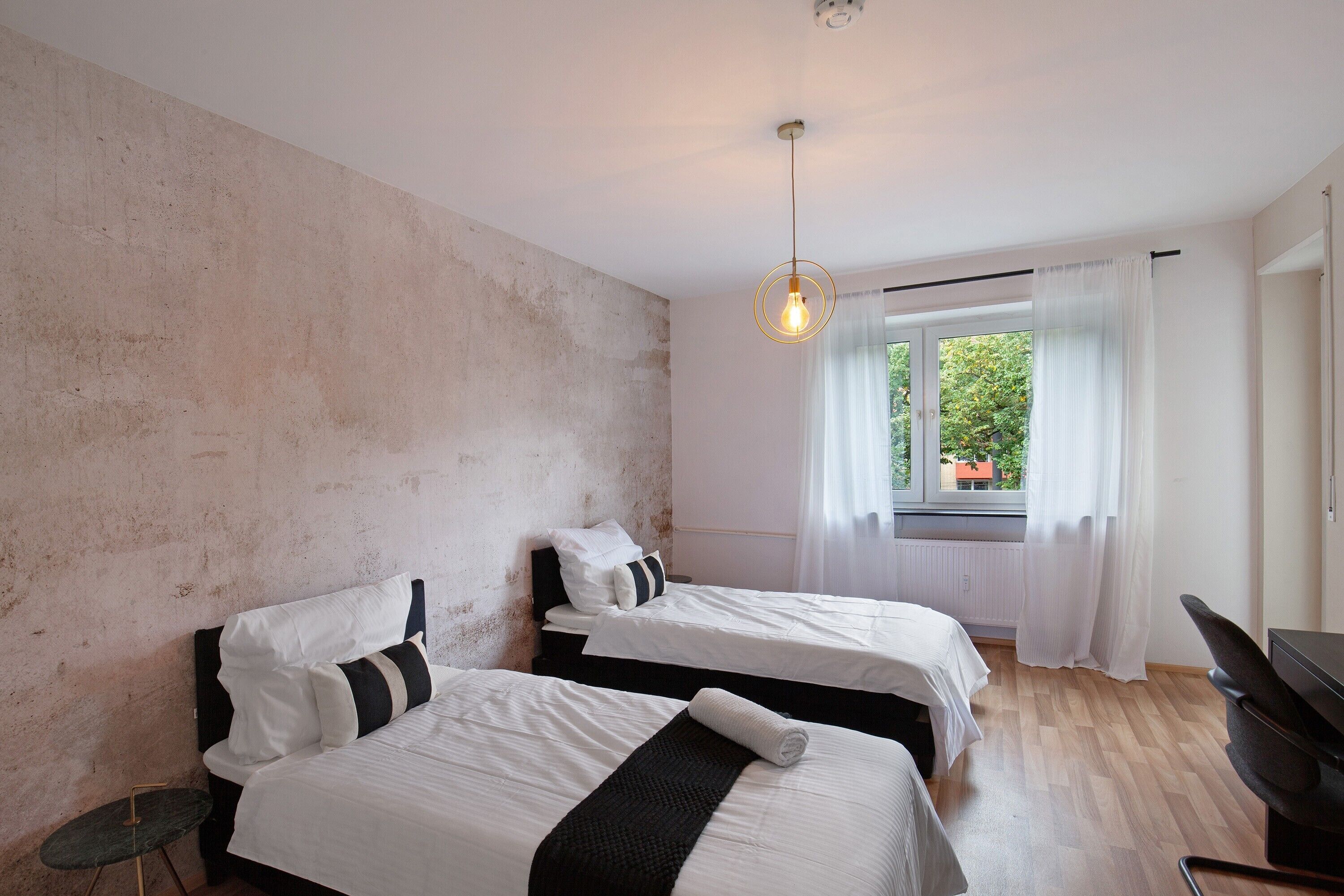 Una habitación con dos camas, una mesita, una silla y una ventana con cortinas.