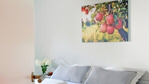 Ein Schlafzimmer mit einem Bett, Kissen, einem Nachttisch, einer Vase mit Blumen und einem an der Wand hängenden Bild von Äpfeln.