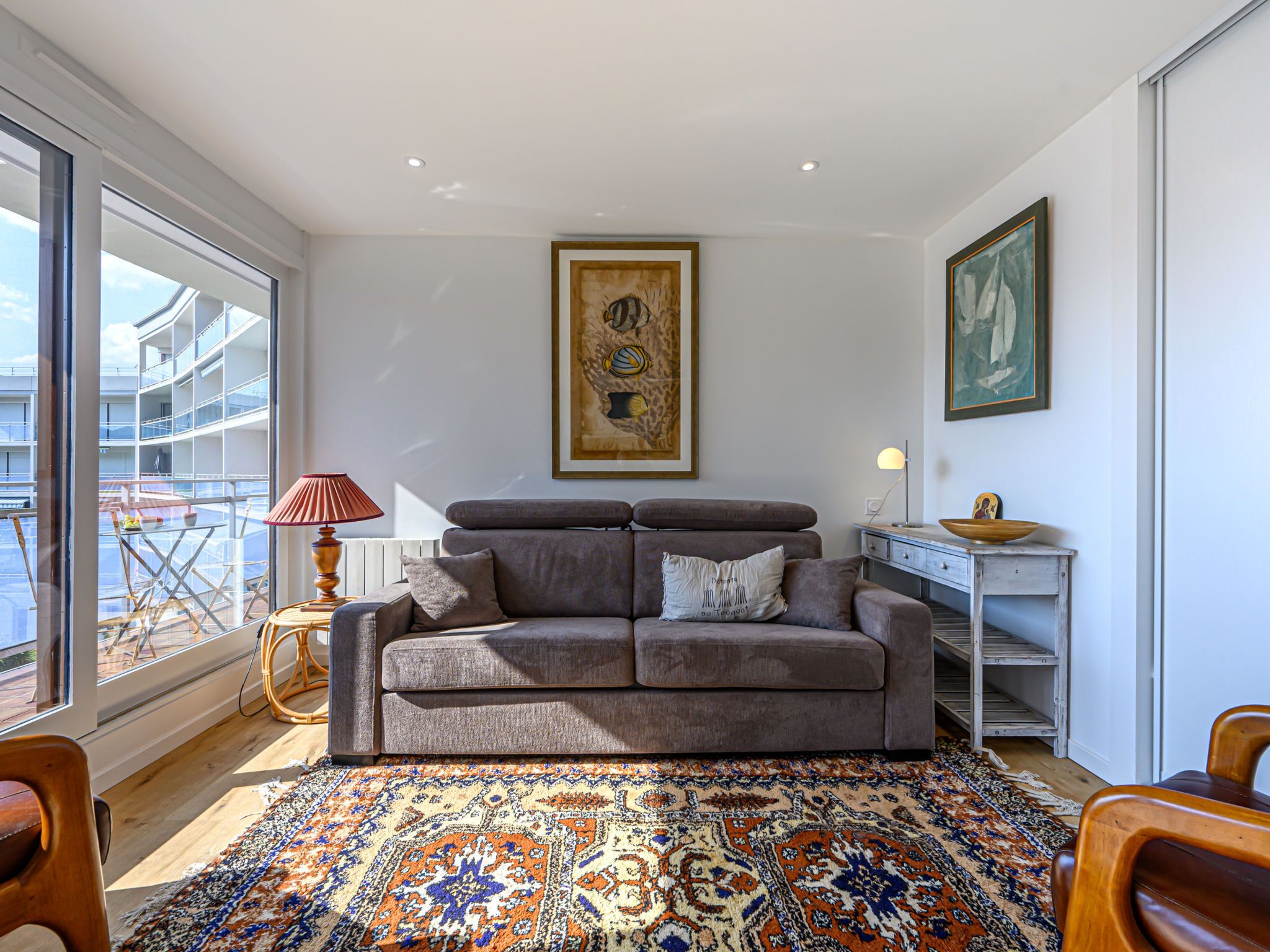 A modern living room with a grey sofa, a patterned rug, and a view of a balcony with outdoor furniture.