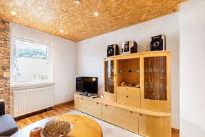 A living room with a wooden ceiling, a stone wall, a window with a view, a TV, a wooden cabinet with glass doors, and a round table with a vase.
