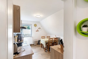 A compact kitchen with a dining area, wooden cabinets, a window, and a green shelf with a plant.