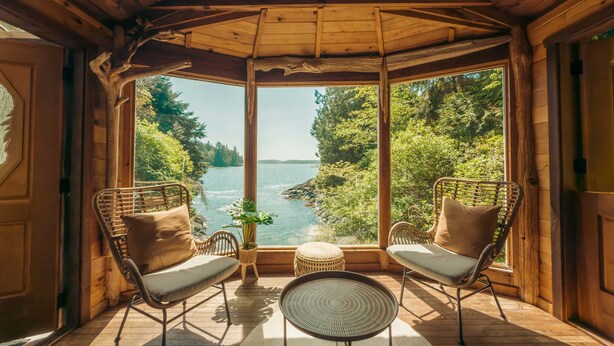 Une cabane en bois dotée d’une véranda en verre, deux fauteuils en osier, une petite table et une vue sur un lac entouré d’arbres.