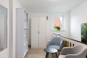 A room with a white wardrobe, a grey armchair, a round table, a window with a plant, and a view of greenery outside.
